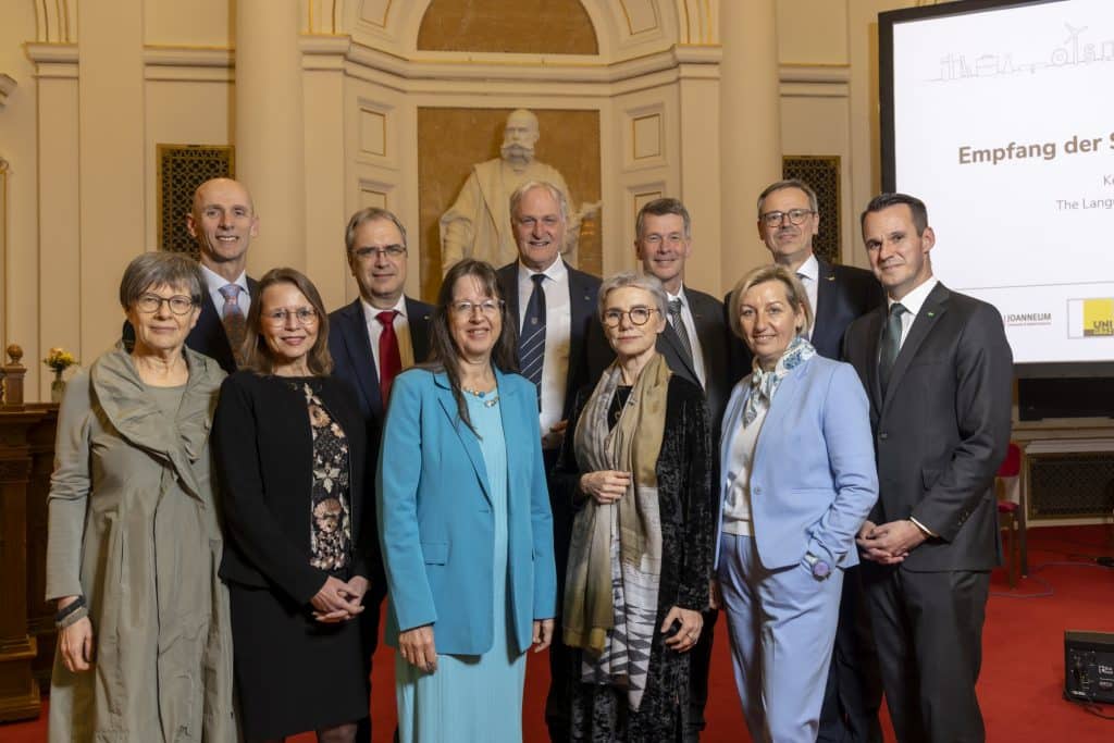 Eine Gruppe elegant gekleideter Personen steht dicht beisammen in einem festlich wirkenden Saal mit hohen, cremefarbenen Wänden und Säulen. Im Hintergrund ist eine große weiße Statue vor einer goldenen Nische zu sehen. Rechts steht ein hoher Bildschirm mit weißer Präsentationsfolie und mehreren Logos im unteren Bereich. Der Boden ist mit rotem Teppich ausgelegt, und die Personen tragen überwiegend Anzüge, Kostüme und festliche Kleider. ©© Helmut Lunghammer