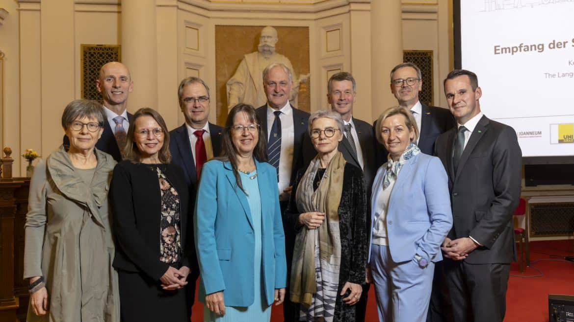 Eine Gruppe elegant gekleideter Personen steht dicht beisammen in einem festlich wirkenden Saal mit hohen, cremefarbenen Wänden und Säulen. Im Hintergrund ist eine große weiße Statue vor einer goldenen Nische zu sehen. Rechts steht ein hoher Bildschirm mit weißer Präsentationsfolie und mehreren Logos im unteren Bereich. Der Boden ist mit rotem Teppich ausgelegt, und die Personen tragen überwiegend Anzüge, Kostüme und festliche Kleider. ©© Helmut Lunghammer