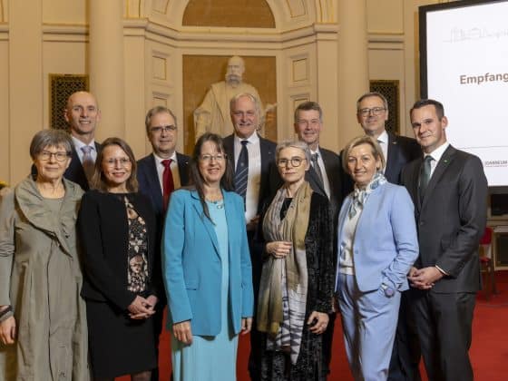 Eine Gruppe elegant gekleideter Personen steht dicht beisammen in einem festlich wirkenden Saal mit hohen, cremefarbenen Wänden und Säulen. Im Hintergrund ist eine große weiße Statue vor einer goldenen Nische zu sehen. Rechts steht ein hoher Bildschirm mit weißer Präsentationsfolie und mehreren Logos im unteren Bereich. Der Boden ist mit rotem Teppich ausgelegt, und die Personen tragen überwiegend Anzüge, Kostüme und festliche Kleider. ©© Helmut Lunghammer